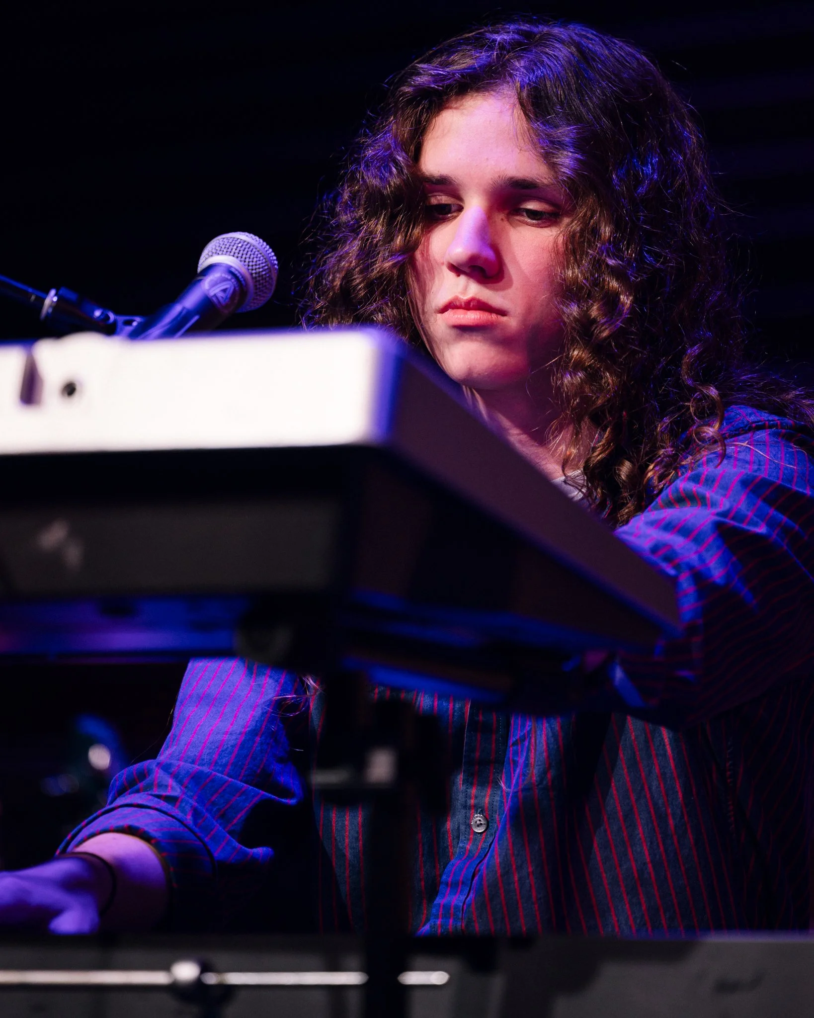 A young woman with curly hair focused on playing a keyboard on stage, with a microphone nearby and stage lighting.