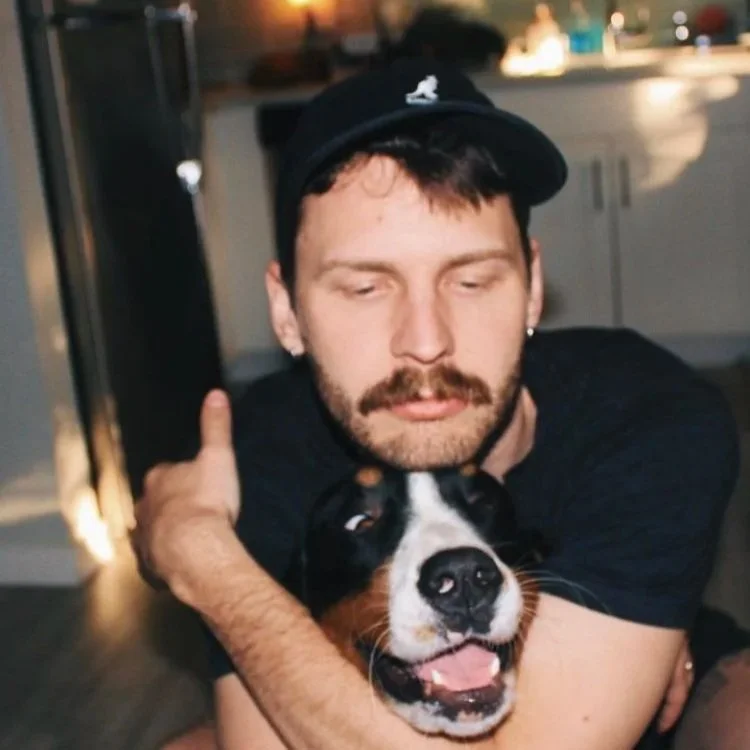 A man with a beard, mustache, earrings, and a black cap hugging a black and white dog with a happy open mouth inside a room.