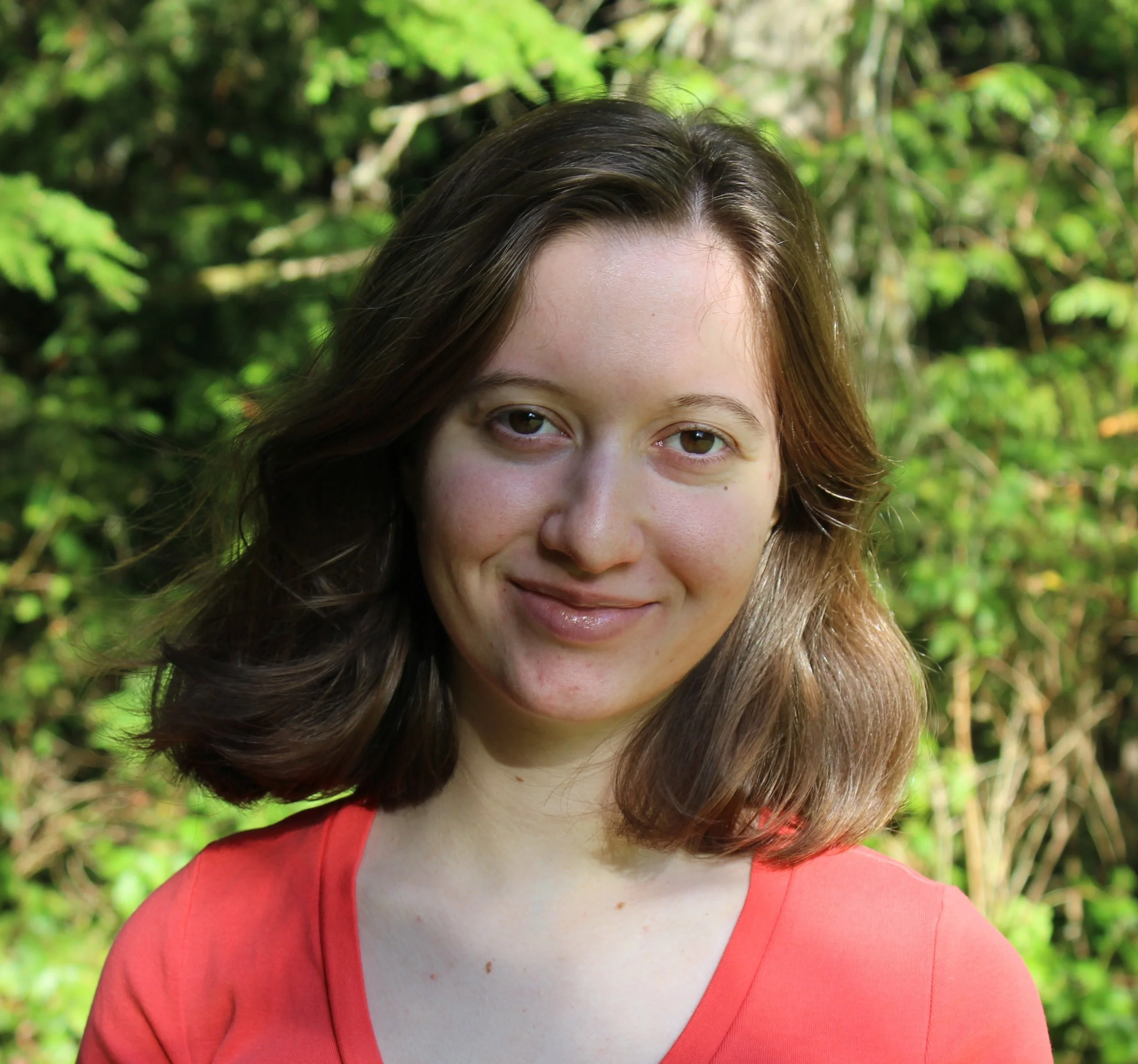 Close-up of a woman's face with brown hair, outdoors with green foliage in the background, wearing a red top.