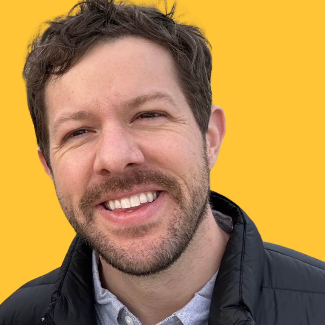 A smiling man with brown hair and a beard, wearing a dark jacket and a light-colored shirt, against a yellow background.