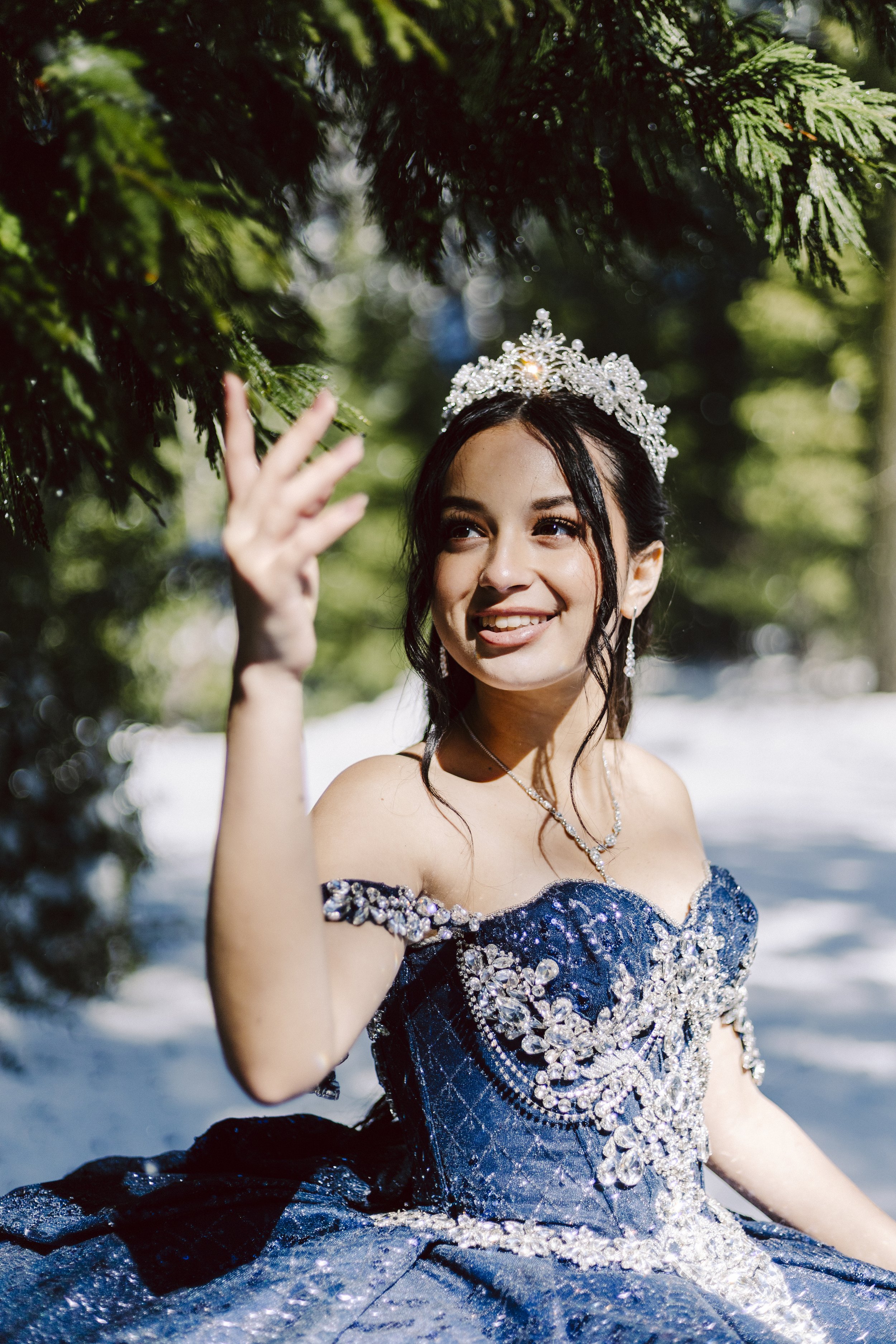 Smiling quinceañera in a blue gown gently touching a snow-covered tree during a winter outdoor photoshoot.