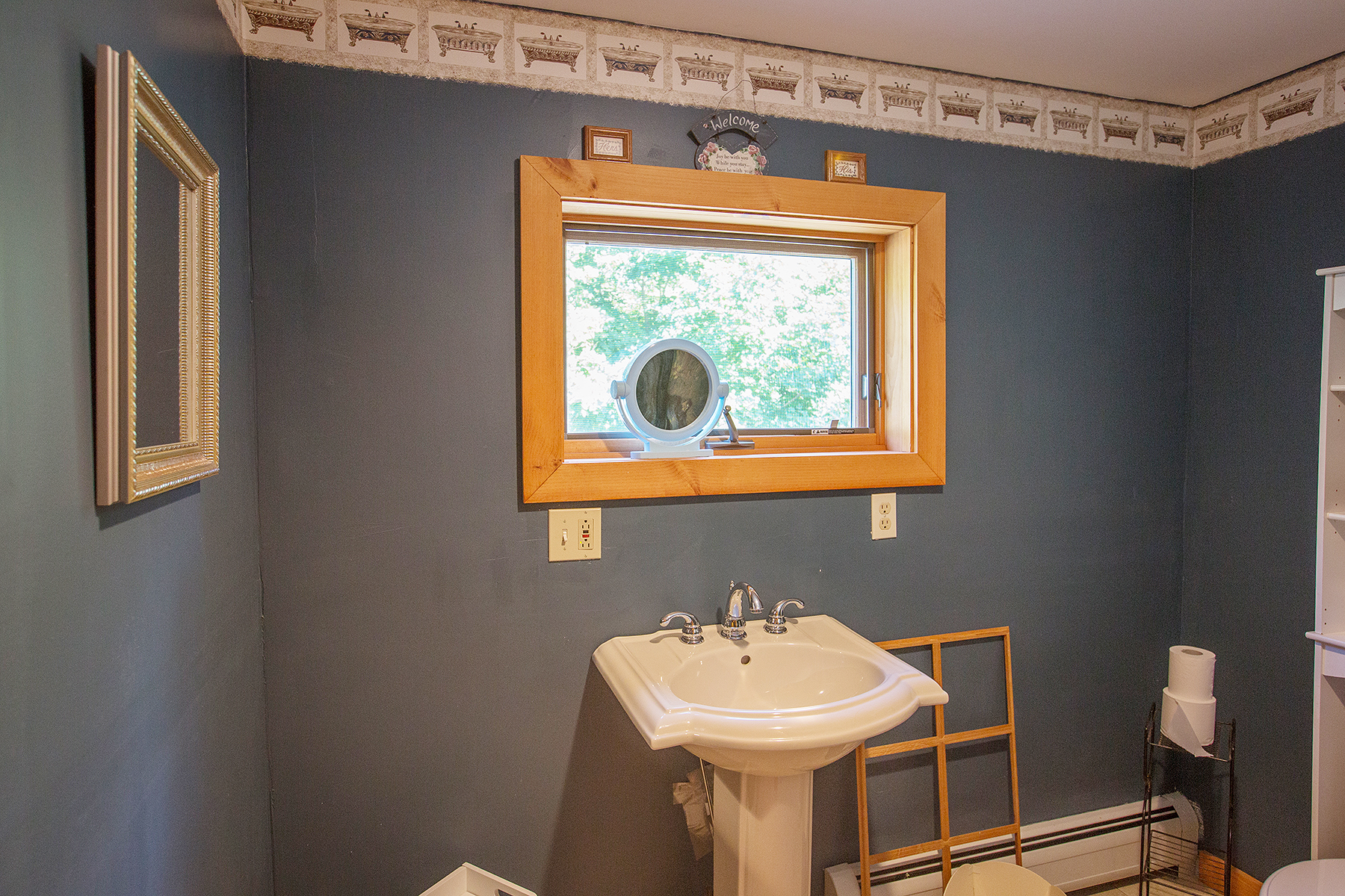  Bathroom with pedestal sink and dark grey walls.  Small window is above the sink that has a wood frame around it. 