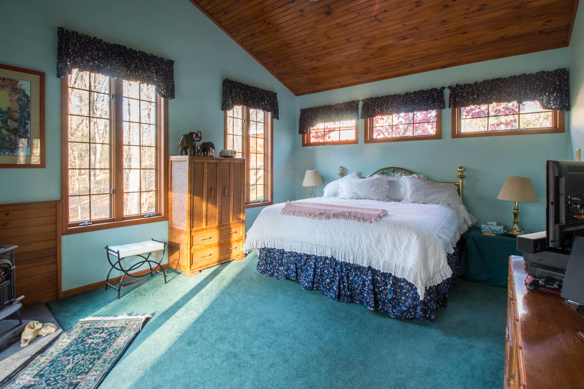  Bedroom with wood bead board ceiling, bluish green carpet and a lot of natural light from windows that outline the walls. 