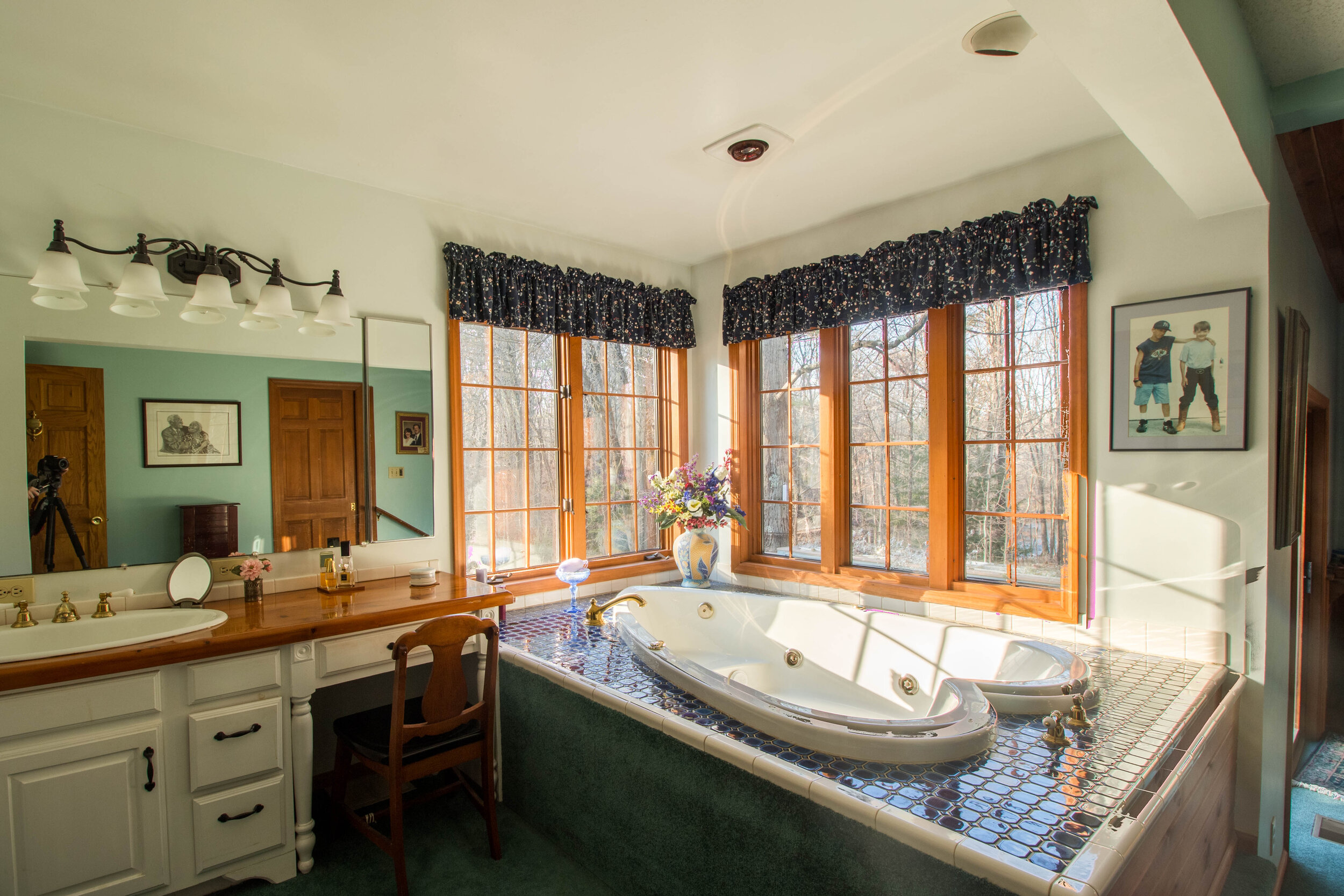  Bathroom with jacuzzi type tub.  Large white vanity with wood top runs alongside the back wall. Rectangle mirror above the vanity/sink combo and a set of 5 windows surround the tub. 