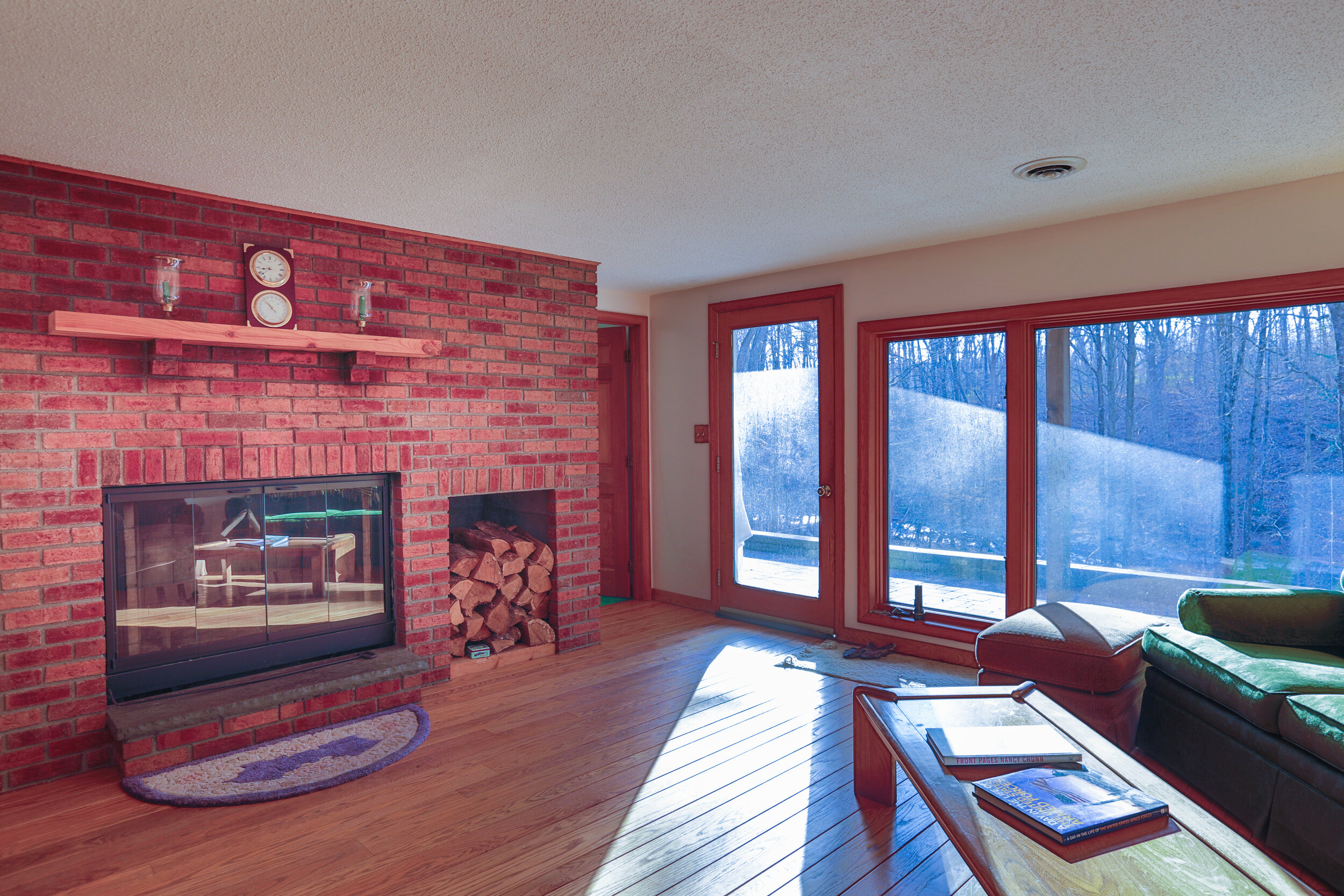  Living room with brick fire place.  Fire is not lit in the fireplace.  Floating wood shelf is hanging above the fireplace with decorations on them.  To the right of the photo is a row of floor to ceiling windows looking outside. 