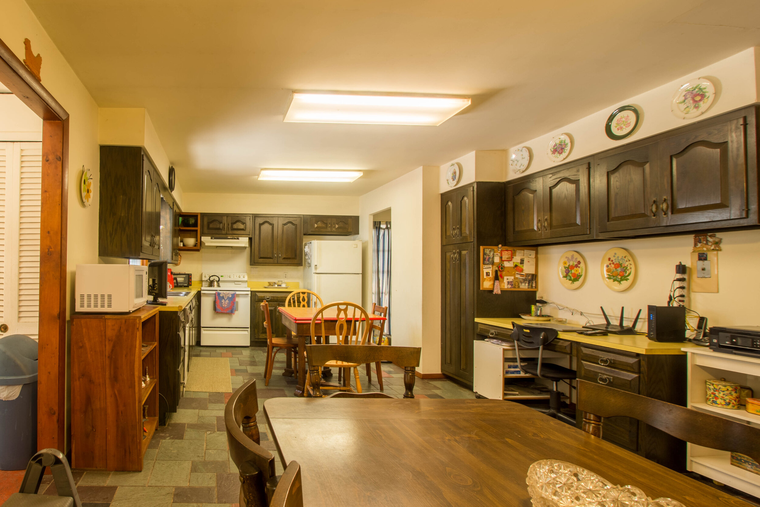  Kitchen with white electric stove and dark wood cabinets.  Two tables are int eh center of the kitchen with shelves around it. 