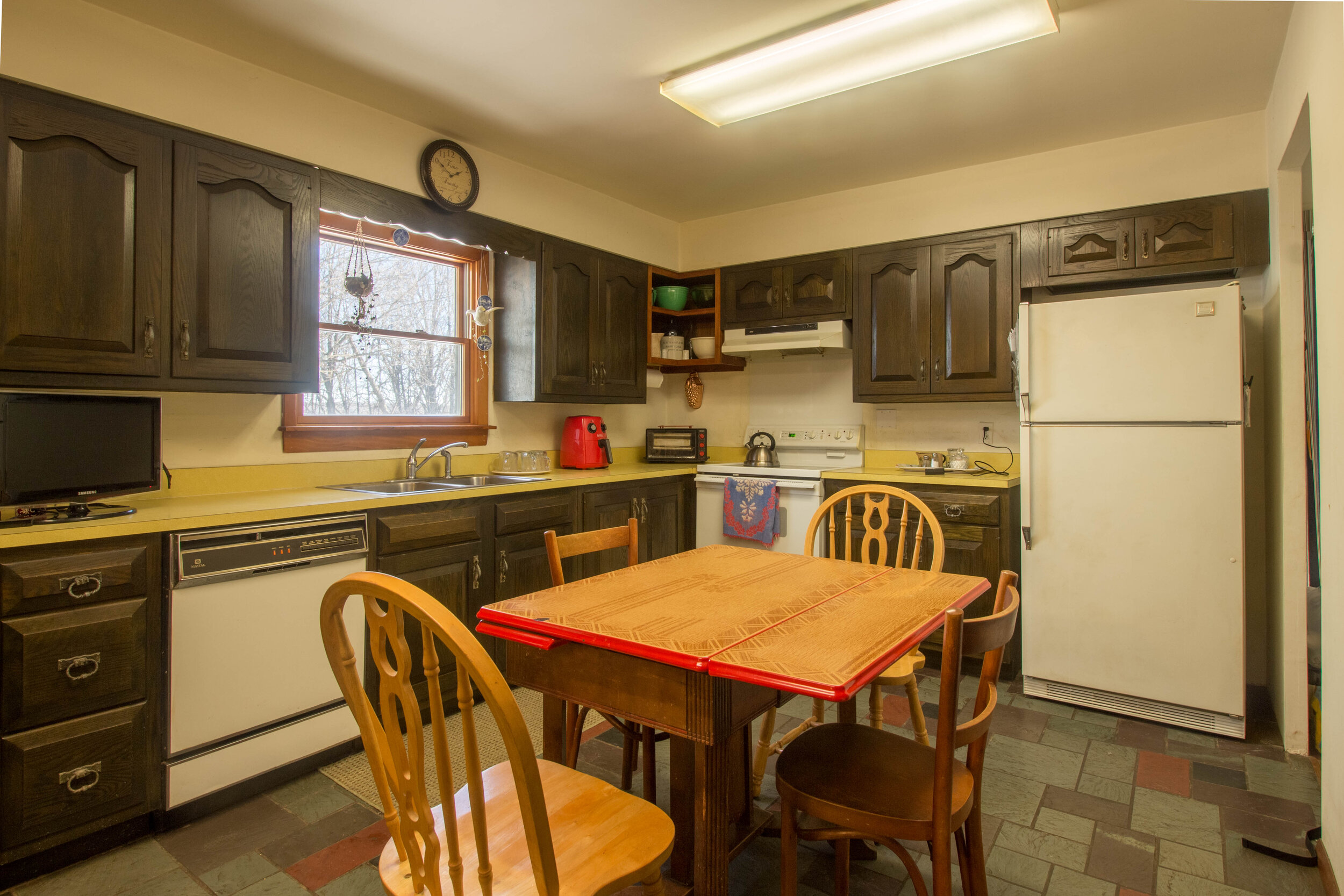  Kitchen with smaller light wood table in the center.  White appliances and dark wood cabinets.  Yellowish countertops with window above the sink. 