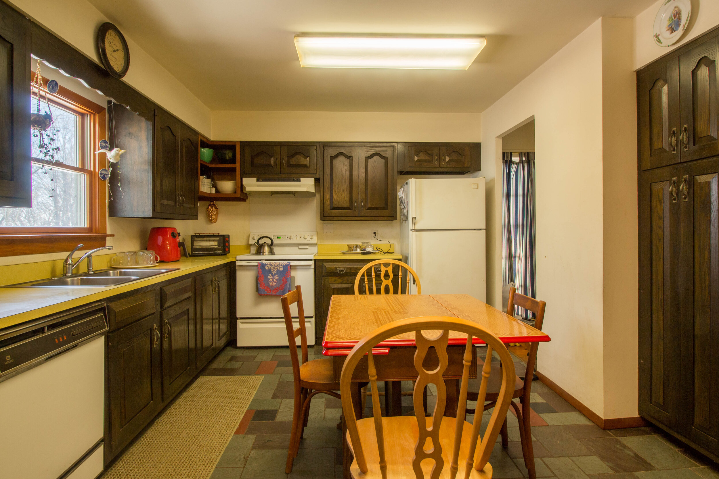  Kitchen with smaller light wood table in the center. White appliances and dark wood cabinets. Yellowish countertops with window above the sink. 