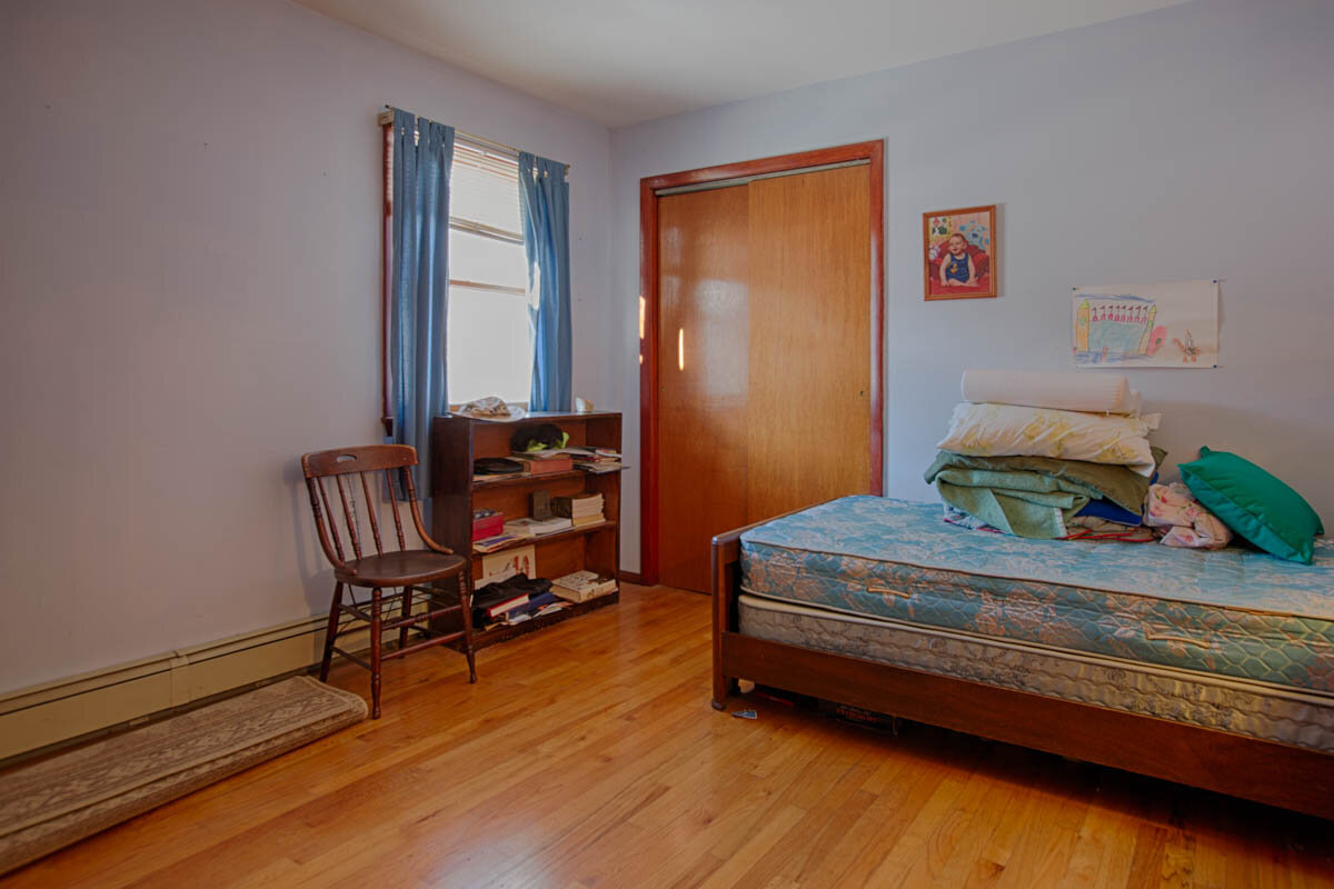  Bedroom with hardwood floors, sliding closet door and a bed resting against the wall.  Single window with blue curtains and a little book shelf in front of the window with a wood chair next to it. 