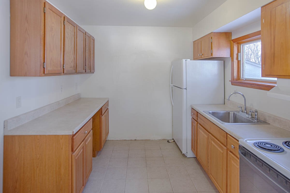  Galley kitchen with wood cabinets and beige countertops.  White refrigerator and electric stove. 