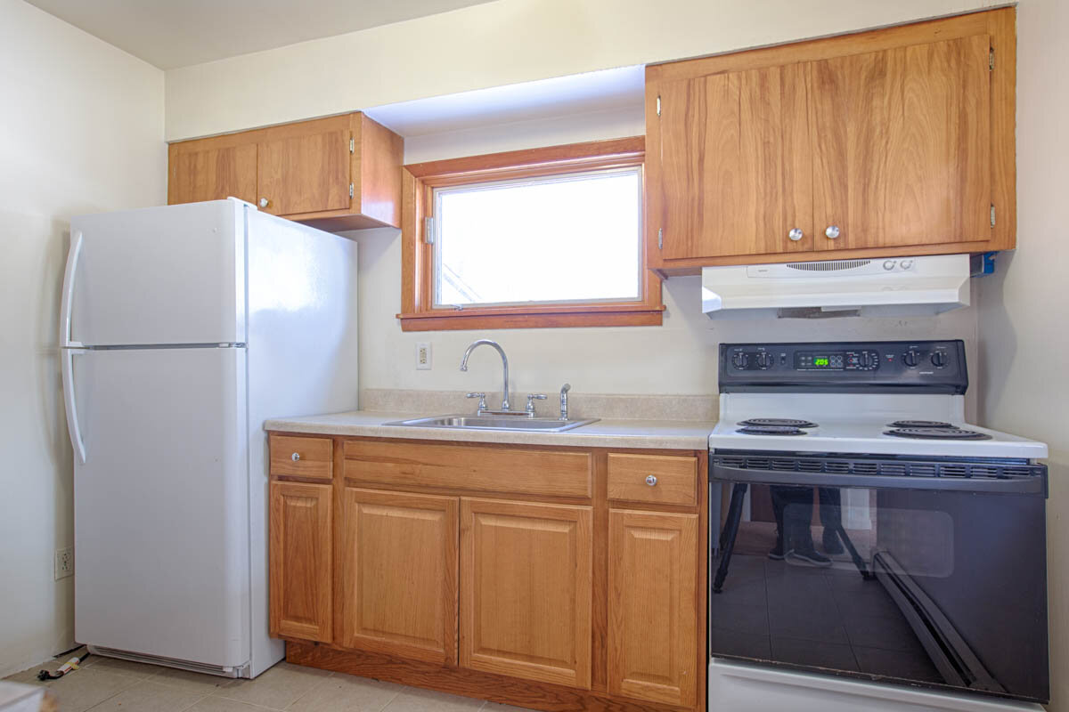  Kitchen with white refrigerator next to a counter with sink in the middle and stove to the right.  Cabinets above the stove and refrigerator. 