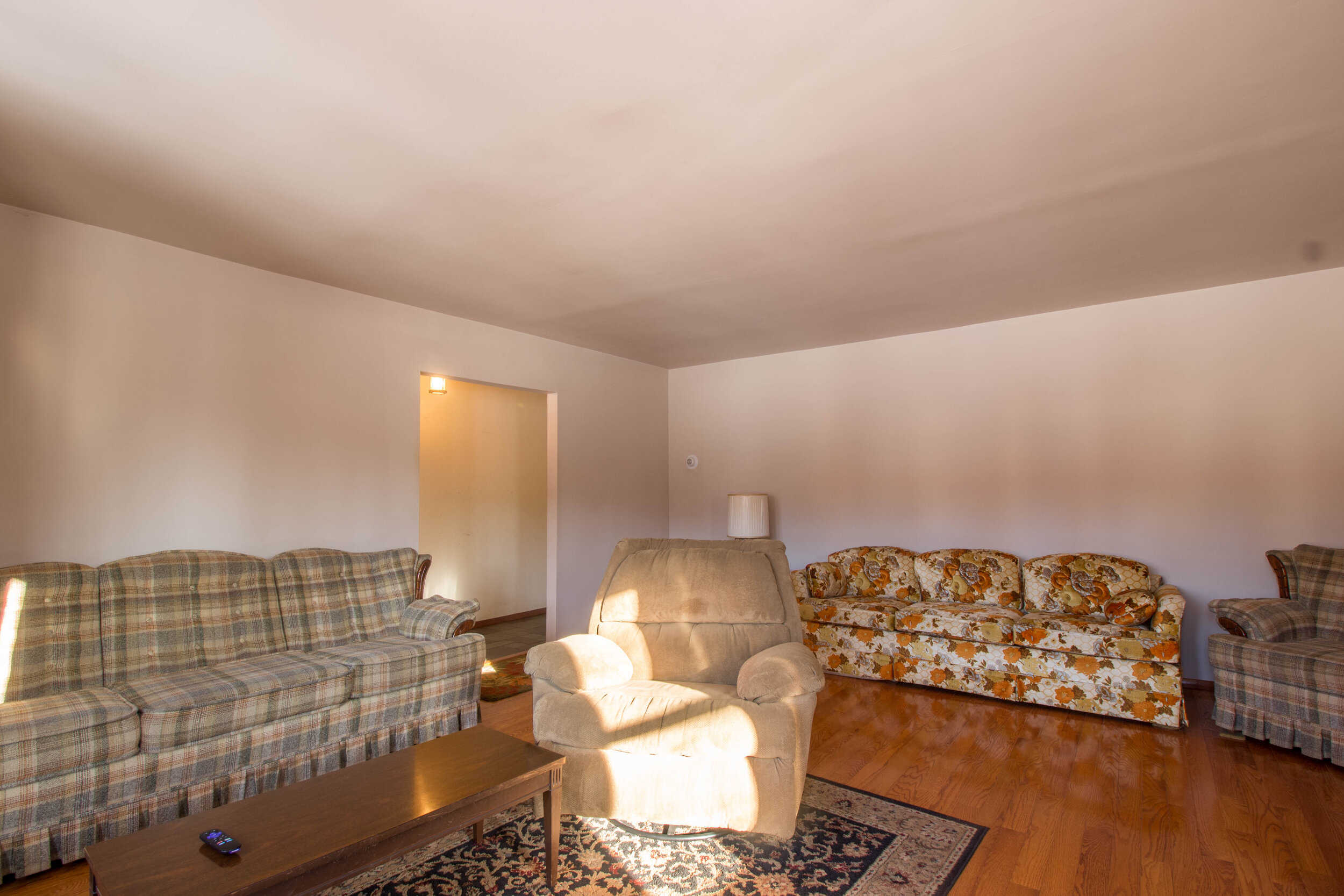  Plaid couch on the left wall, floral couch on the center wall and a recliner in the center on an area rug. Hardwood floors are refinished and glossy looking.  