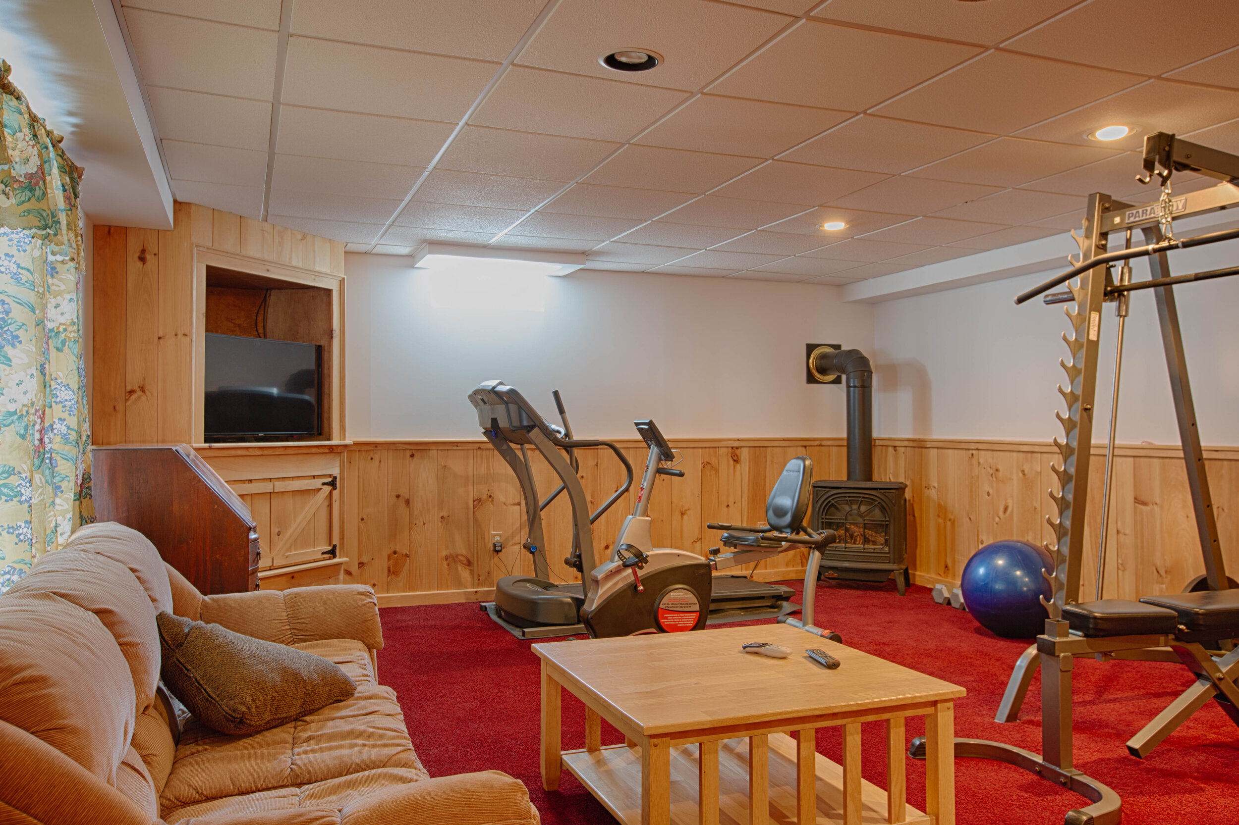  Basement with couch to the left, table in the center and workout equipment in the background on top of a red rug.  Also, there is a wood burning stove in the corner of the photo. 