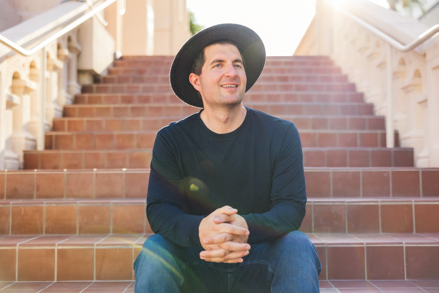 man wearing hat sits on steps and smiles while sun flares in