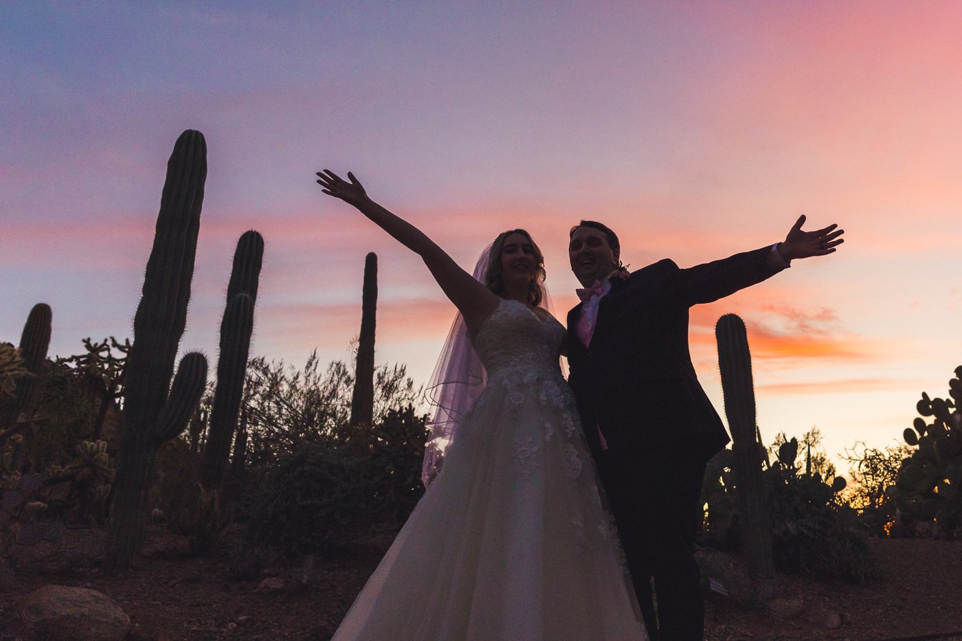 bride and groom silhouetted at sunset throw arms in the air
