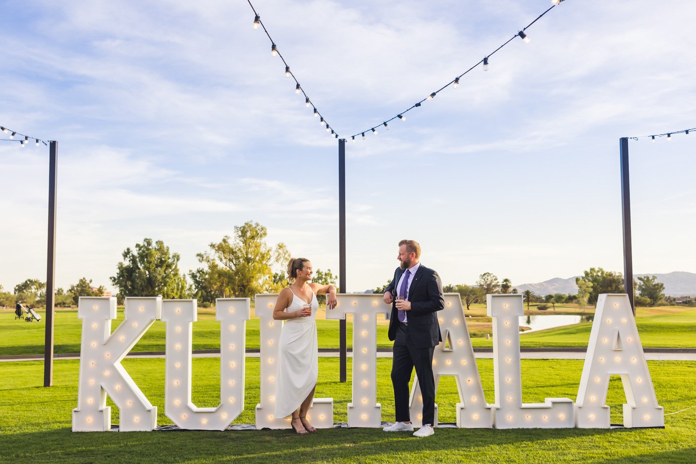 bride and groom holding drinks in front of their name sign at wedding