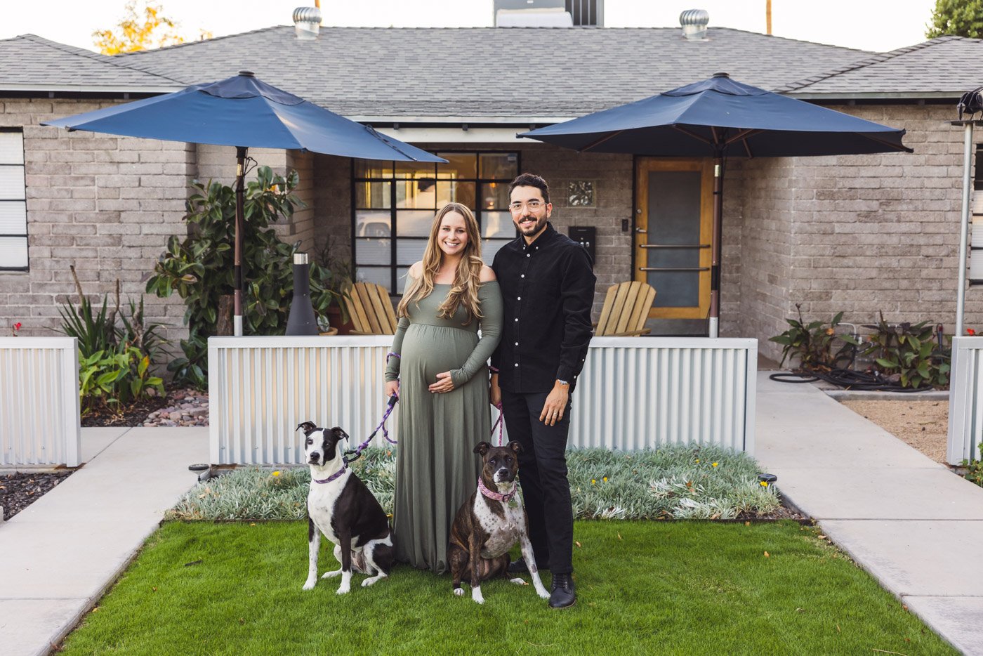 expecting couple with their dogs in front of their gray brick house