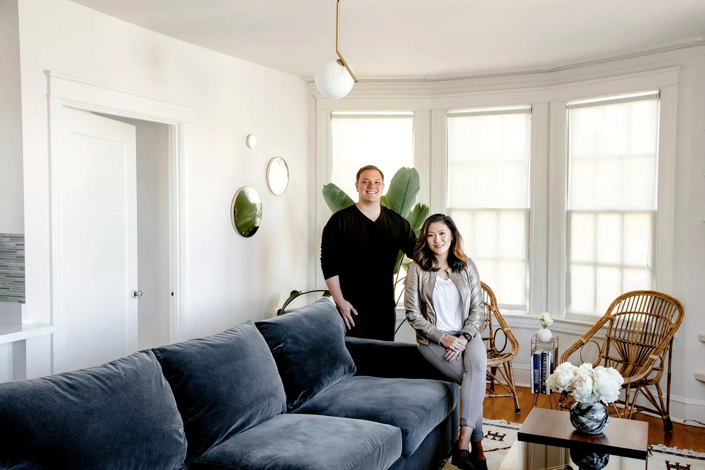 Niles Lichtenstein and Alicia Cheung Lichtenstein at home. When the couple got together, they opened a joint account to use for redecorating the apartment. The sofa is from Crate & Barrel , the rattan chairs are from Blue Ocean Traders , the coffee table is vintage and from Donny O Antiques . The Moon mirrors are by artist Heather Palmer and the 1C pendant from Flos by Michael Anastassiades.