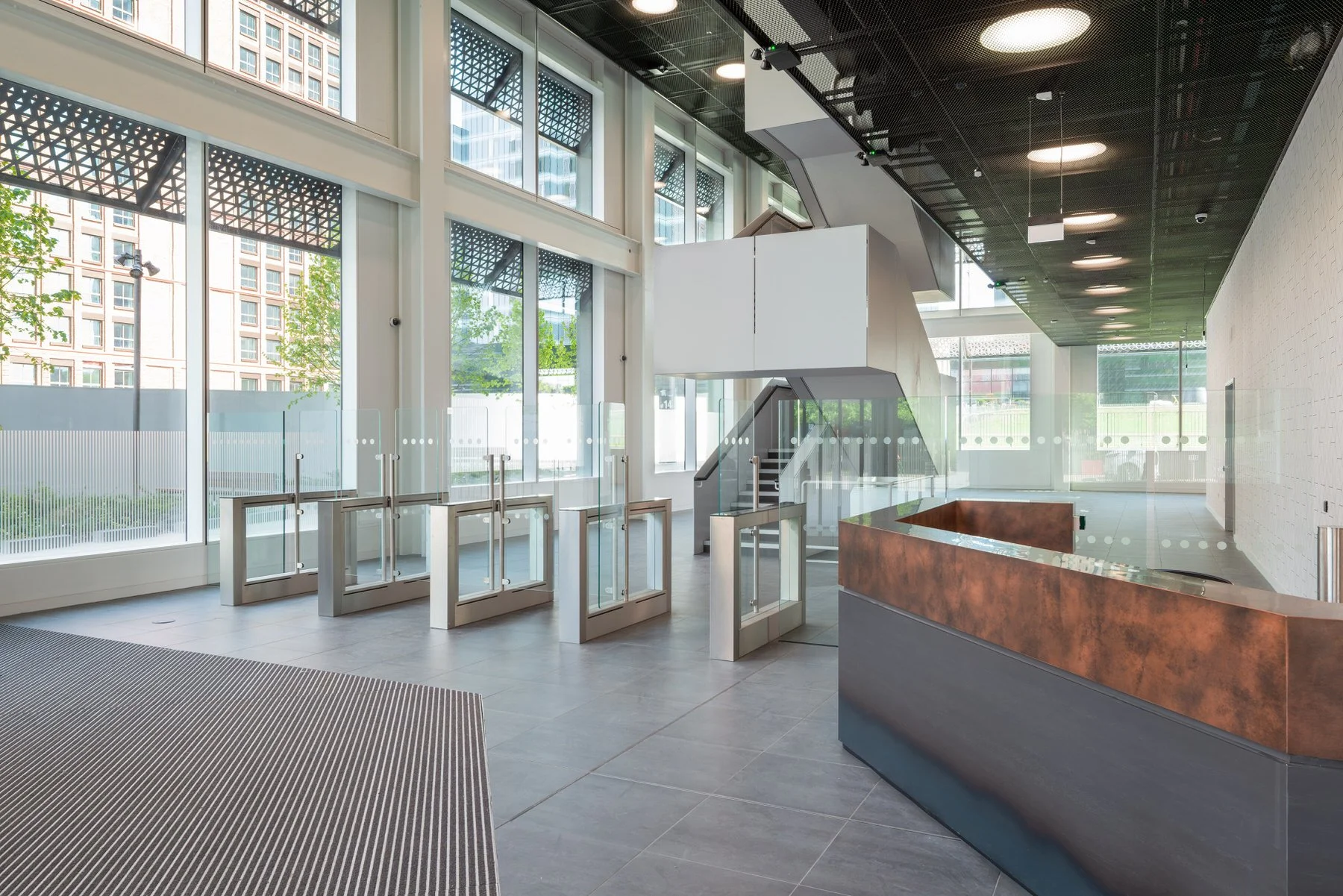 Modern office lobby with large windows, metal security gates, a reception desk, and a staircase leading to an upper level.