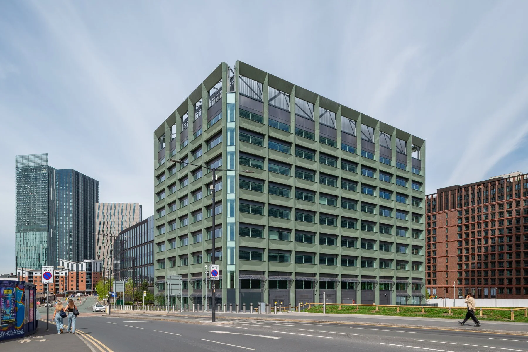 Modern multi-story building with a grid-like facade, situated on a city street with pedestrians walking along the sidewalk and cars parked nearby.