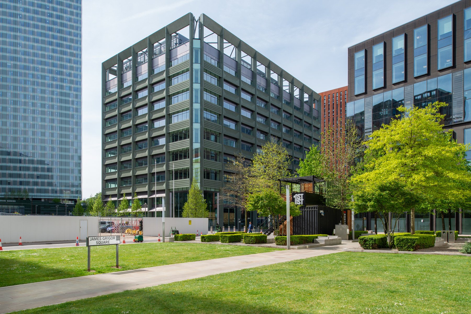A modern cityscape featuring tall office buildings with glass and metal exteriors, green trees, manicured bushes, and a grassy park area in the foreground. There is a sign reading 'JAMES GRIGOR SQUARE' and a small construction site with orange cones.