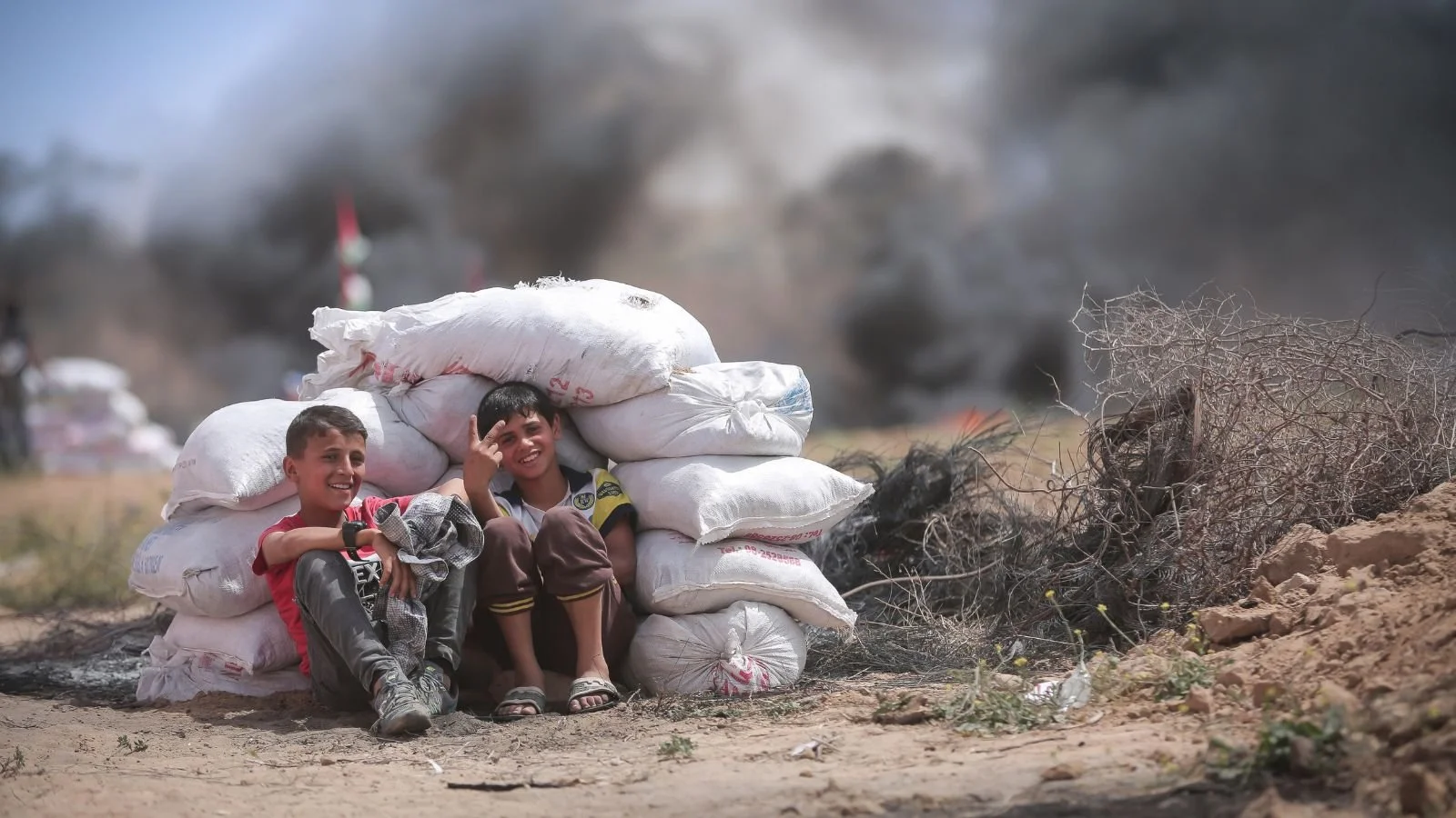 Two smiling boys sitting behind stacked white sacks of supplies on a dusty ground with smoke and fire in the background.
