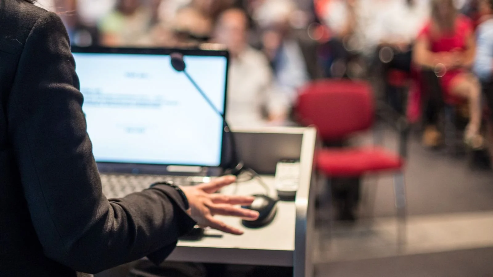 A person in a black jacket using a mouse at a desk with a computer monitor, with a blurred conference room or event setting in the background.