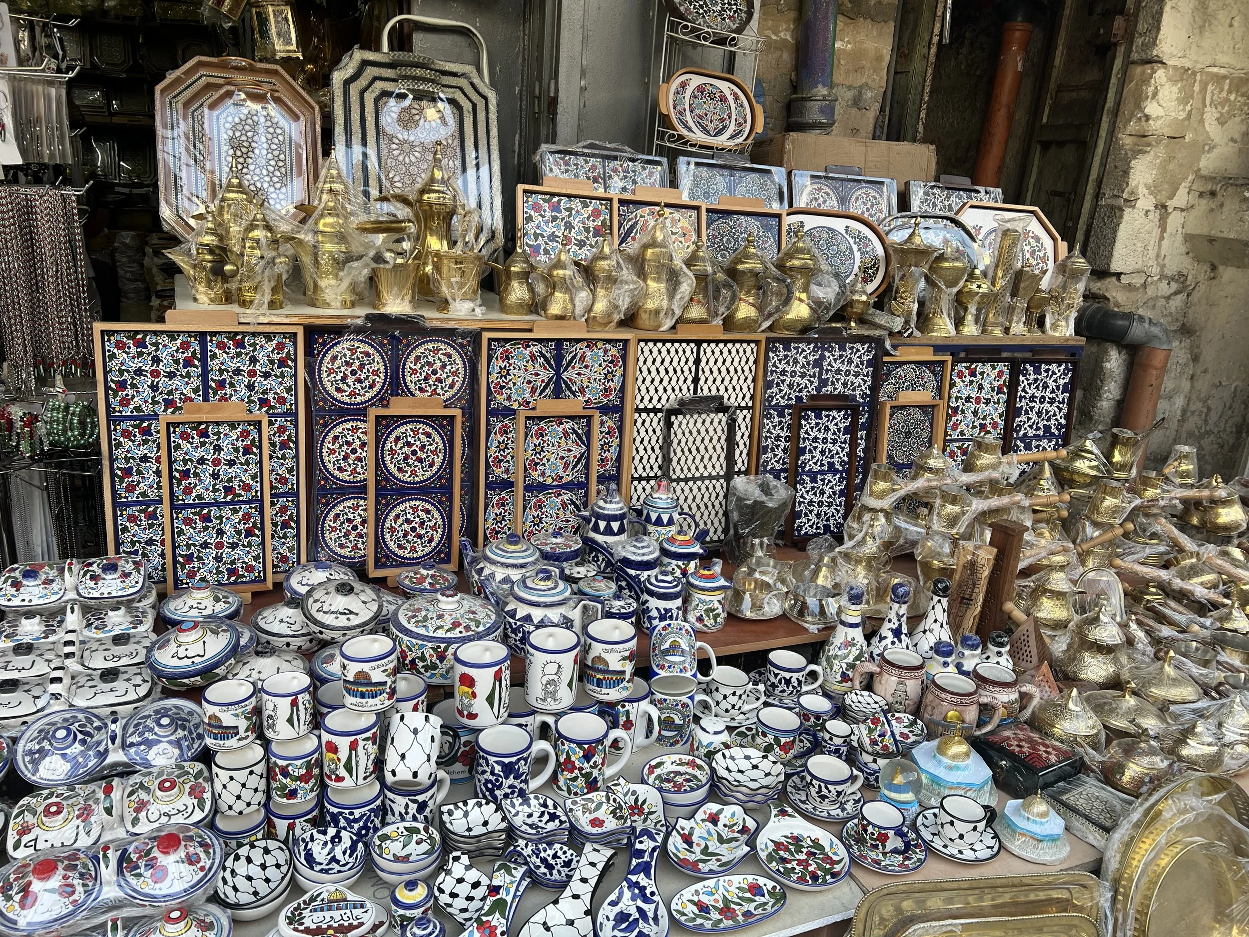 Display of various decorative ceramic and metal items including cups, bowls, and plates, with intricate patterns and colorful designs, arranged on tables at an outdoor market stall.