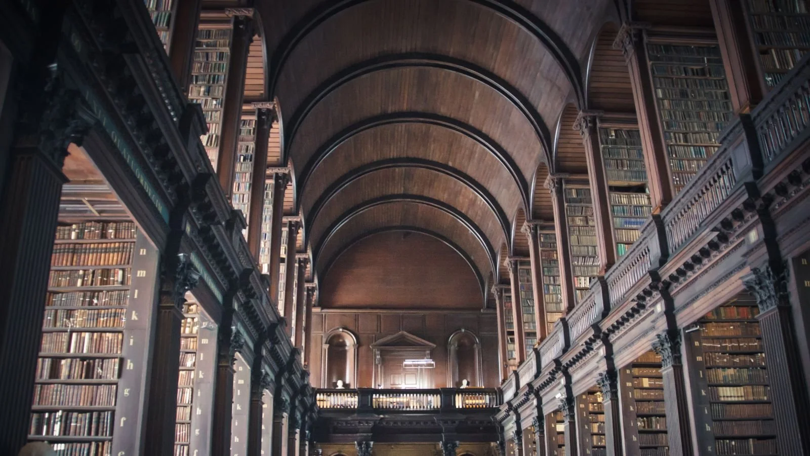 Interior view of a historic library with tall wooden bookshelves filled with books, arched wooden ceilings, and a balcony with ornate railings.