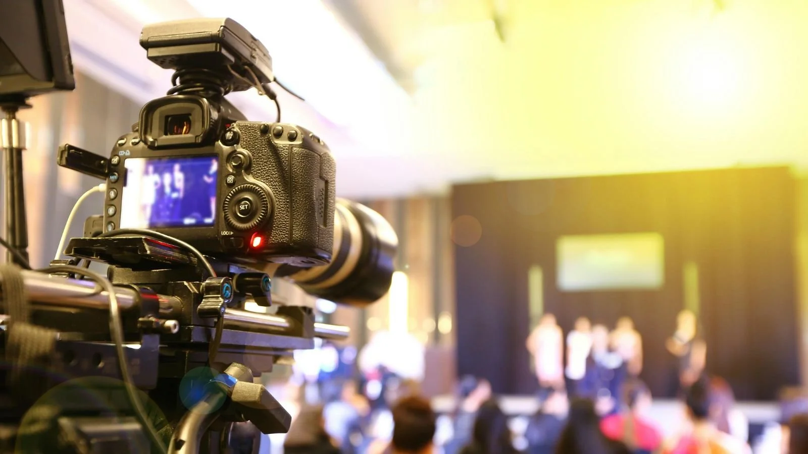 A professional video camera filming a panel discussion or presentation on stage with people standing and a large screen in the background in a brightly lit conference or event hall.