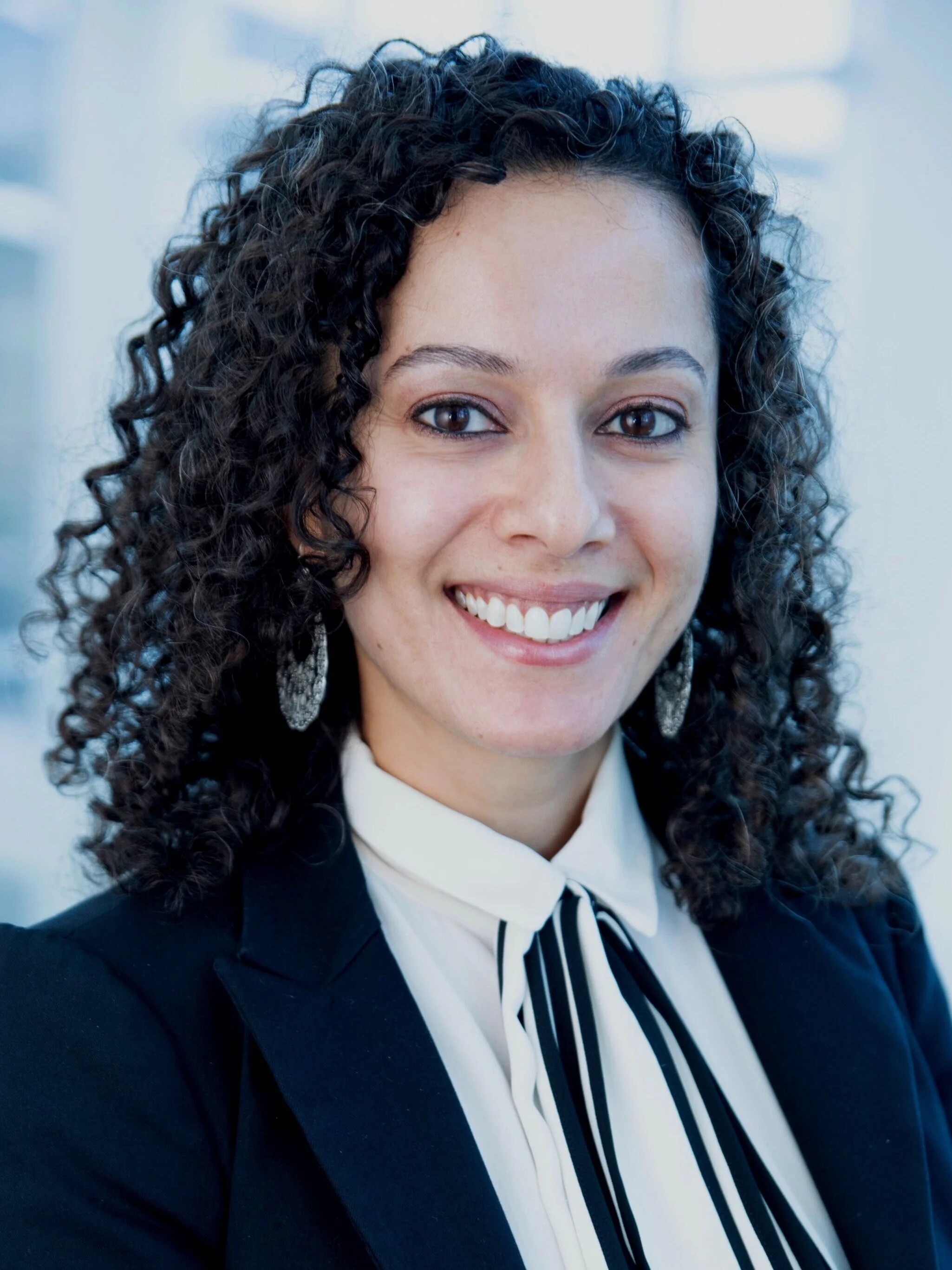 A woman with curly dark hair, smiling, wearing a formal black suit and a white shirt with a black and white striped tie, standing in a bright indoor setting.