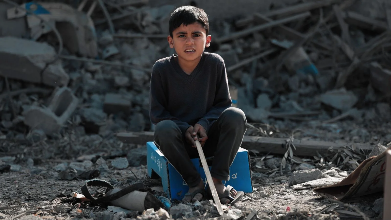 A young boy sitting on a blue box made of cardboard, surrounded by debris and rubble, holding a stick in his hands, with a somber expression.