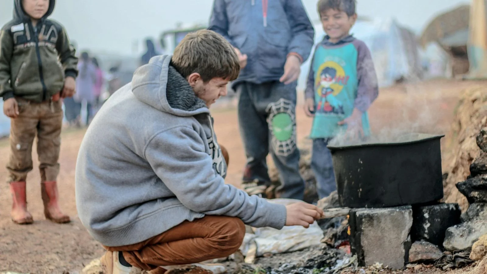 A young man crouches down near a makeshift fire outdoors, tending to a pot on bricks, while three children stand nearby in a camp setting, with tents and vehicles in the background.