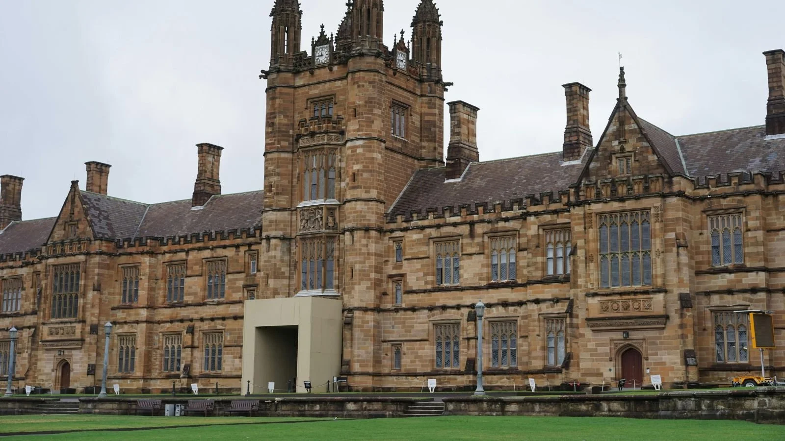A historic Gothic-style building made of brown stone with pointed towers, arched windows, and decorative stonework, set against a cloudy sky, with a well-maintained lawn and benches in the foreground.