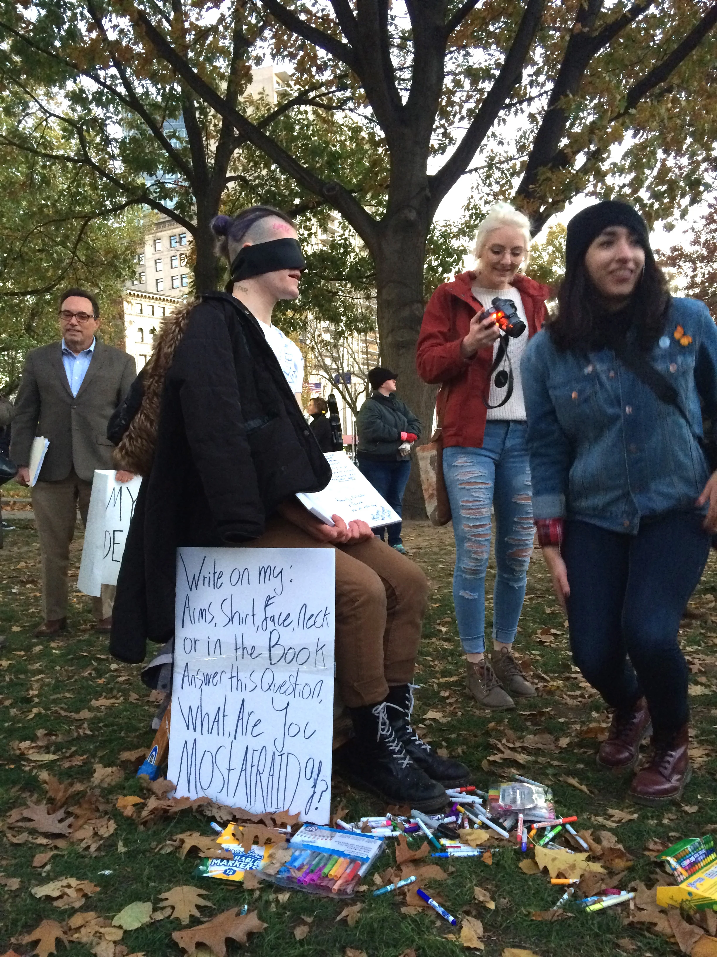 Performance piece. Sign reads: "Write on my arms, shirt, face, neck, or in the book. Answer this question: What are you most afraid of?"