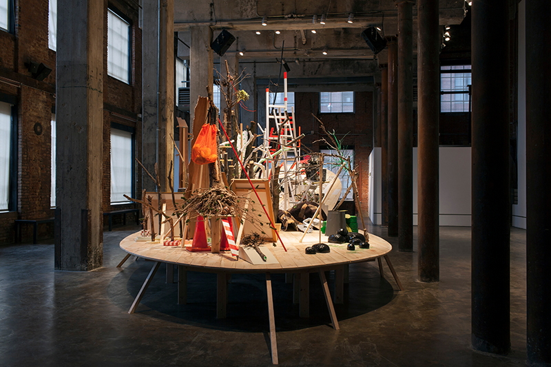Andrea Loefke, Homecoming at Smack Mellon, Brooklyn, 2013. Found tree branches and stumps, wood, ladders, chicken wire, traffic cones, photographs, plaster casts of tree branches, hardware, glitter pigmented latex, pedestals, milk crates, mirrors, carpet, clamps, foam, mixed media, and painted water level on walls
