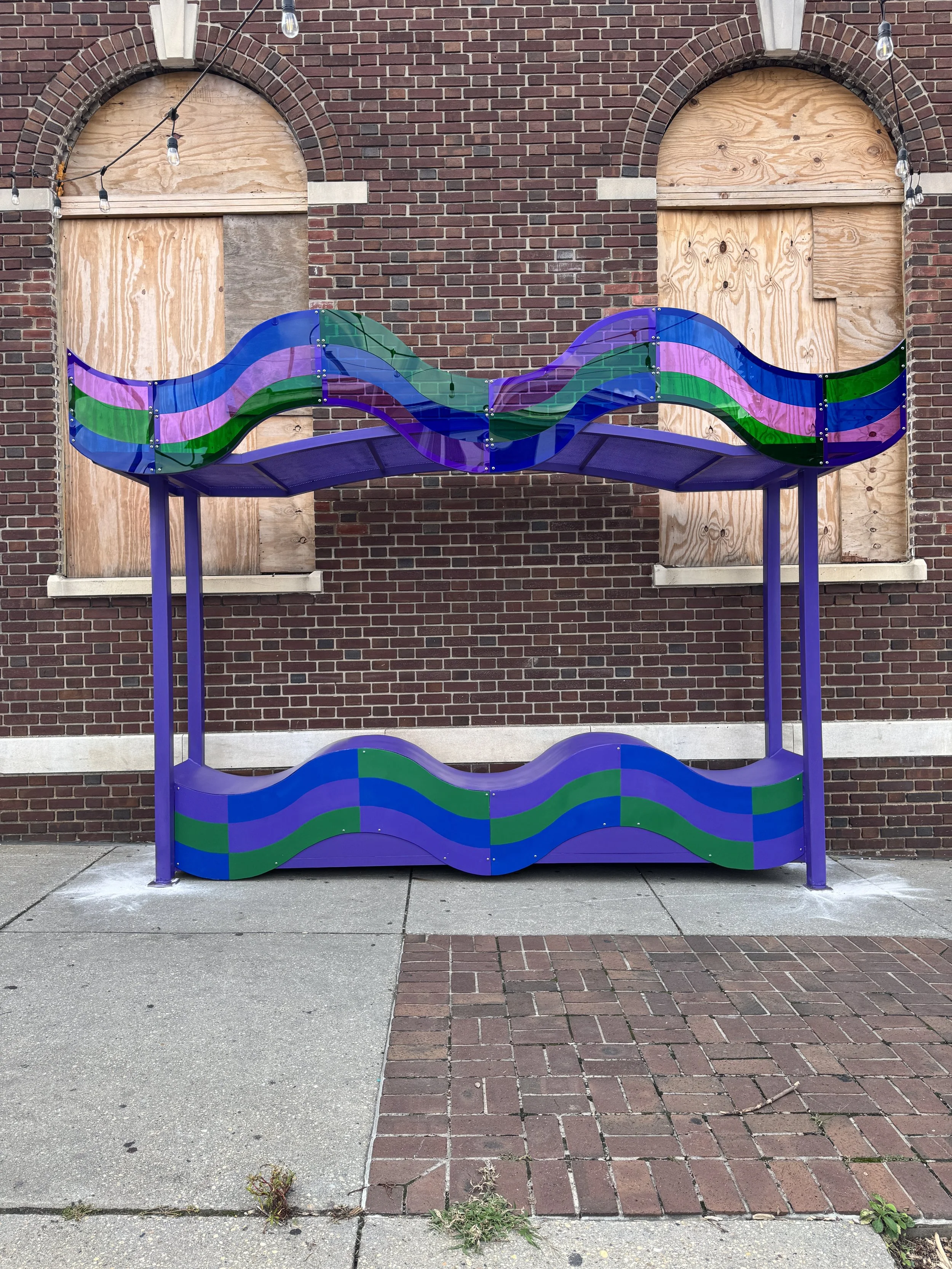 A purple double bench with a wavy base and a canopy of translucent blue, green, and violet bands stands in front of a brick building.