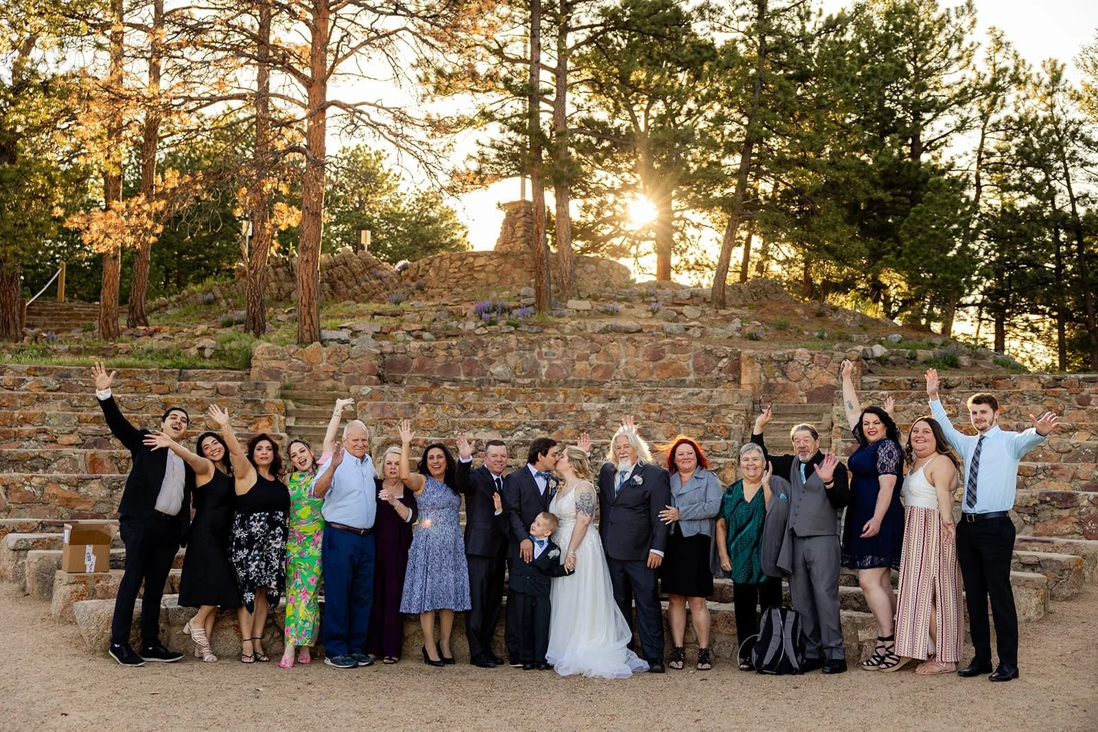 A bride, groom and their 15 guests post happily for a group photo with stone amphitheater seating and setting sun behind them.
