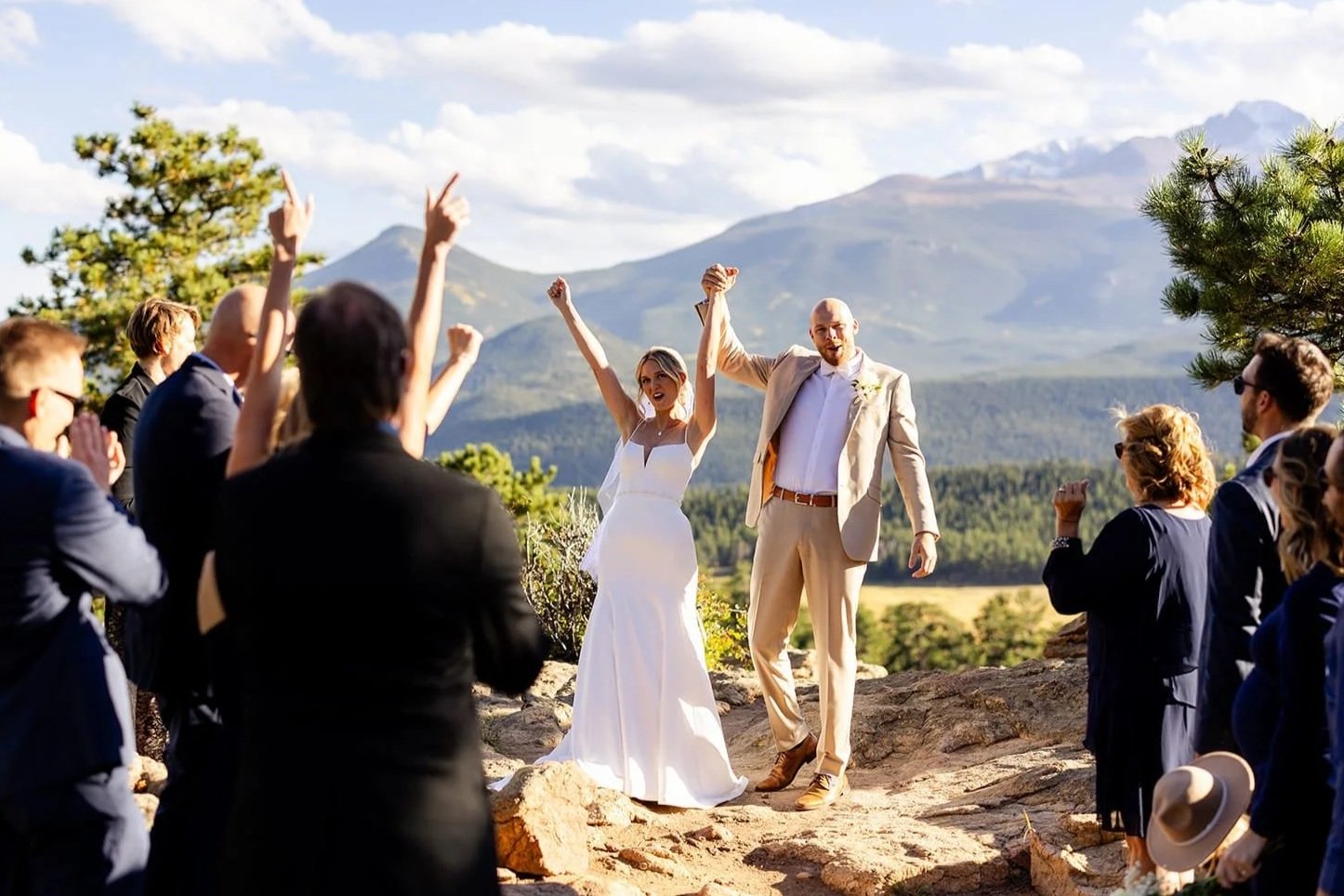 A bride and groom raise their hands in triumph as their elopement ceremony completes, while a small group of guests cheer with a sunset mountain backdrop.