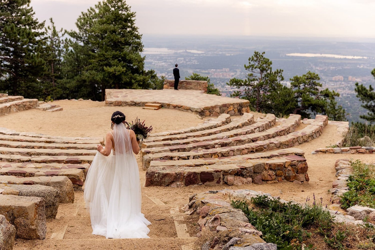 A bride walks a staircase into a stone amphitheater while her groom waits with back turned for a first look.