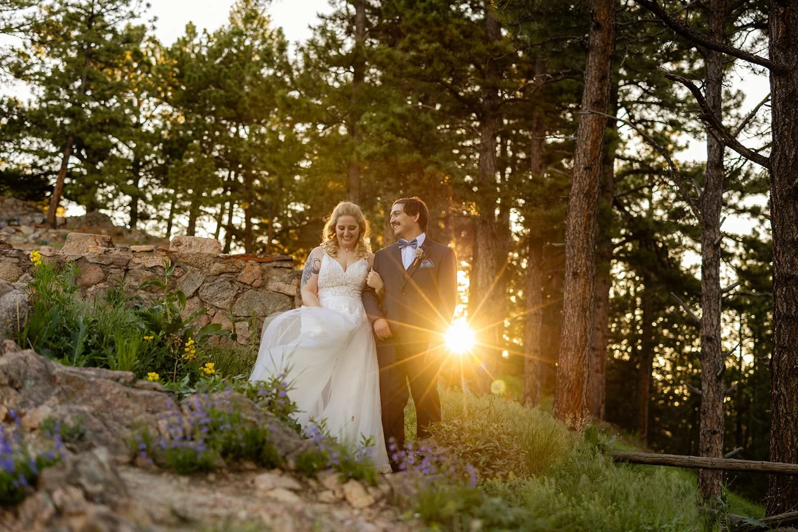 A just-married bride and groom walk arm in arm with a sunset glowing through the trees behind them and yellow and purple wildflowers in the foreground.