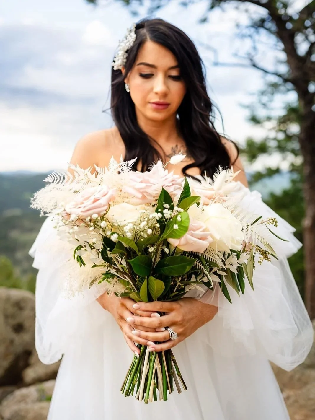 A bride looks down at her bouquet of pink and white roses with feathery embellishments in soft sunset light.
