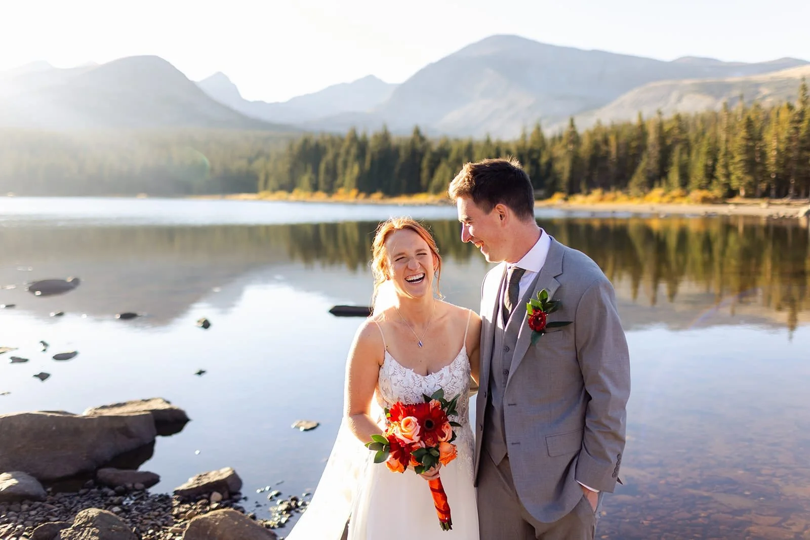 A just-married bride and groom laugh and smile just after their elopement ceremony next to an alpine lake with fall mountain backdrop.