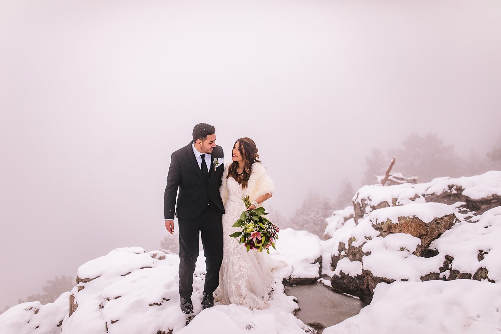 A bride and groom joyfully pose together on a snowy mountain peak, surrounded by a beautiful winter landscape.