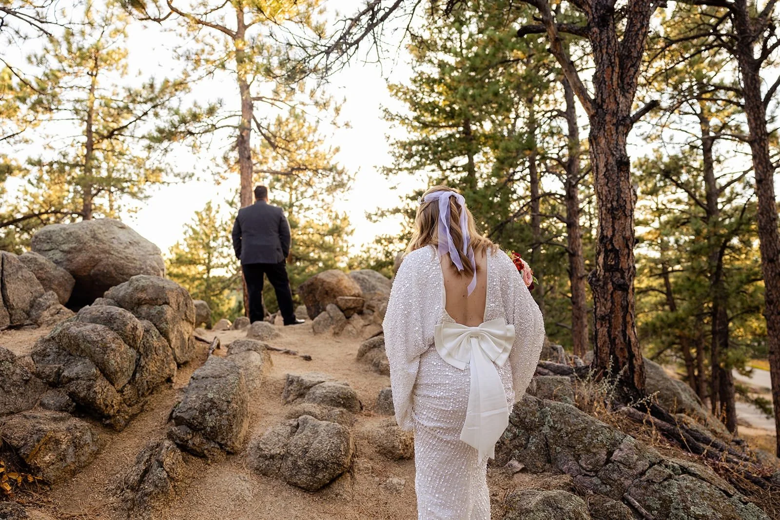 A bride climbs a forested hill to her groom with his back to her so she can surprise him with a first look moment.