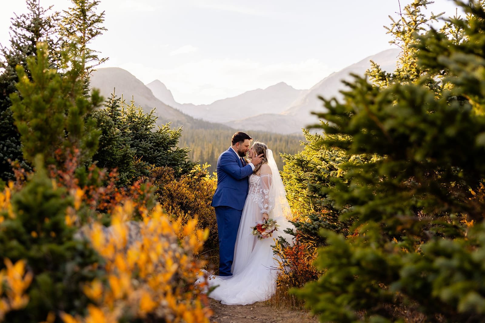 An eloping couple kissing on a mountain path surrounded bushes with fall colors of reds oranges and yellows with peaks in the background.
