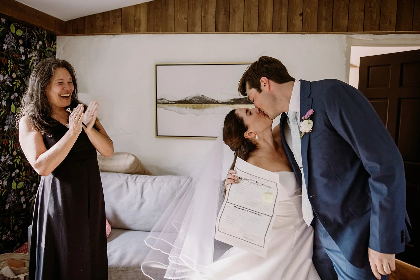 A just-married couple kiss in a living room as the bride holds up their marriage license for the camera and Officiant Lisa claps and smiles.