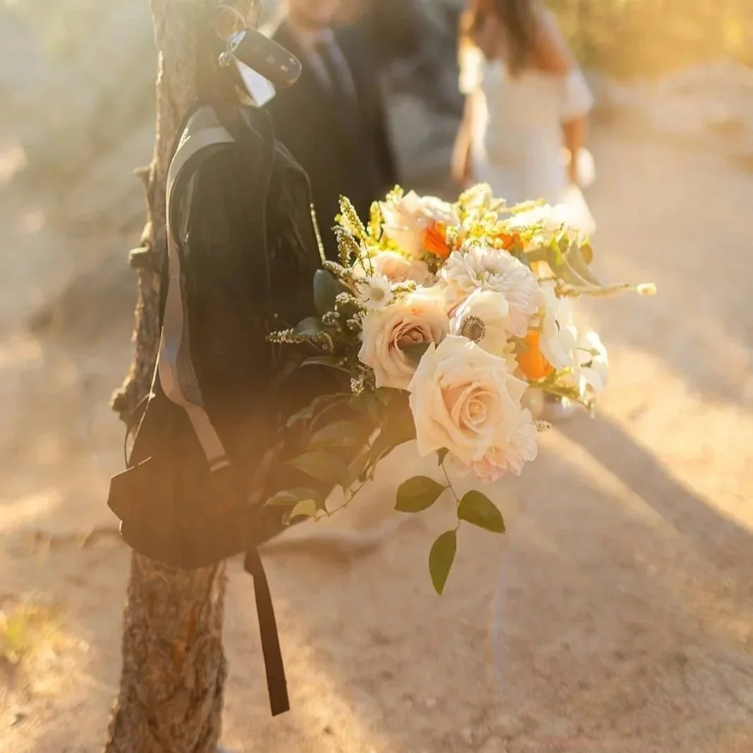 A backpack with a bridal bouquet in it hangs from a small tree trunk in the setting sun.