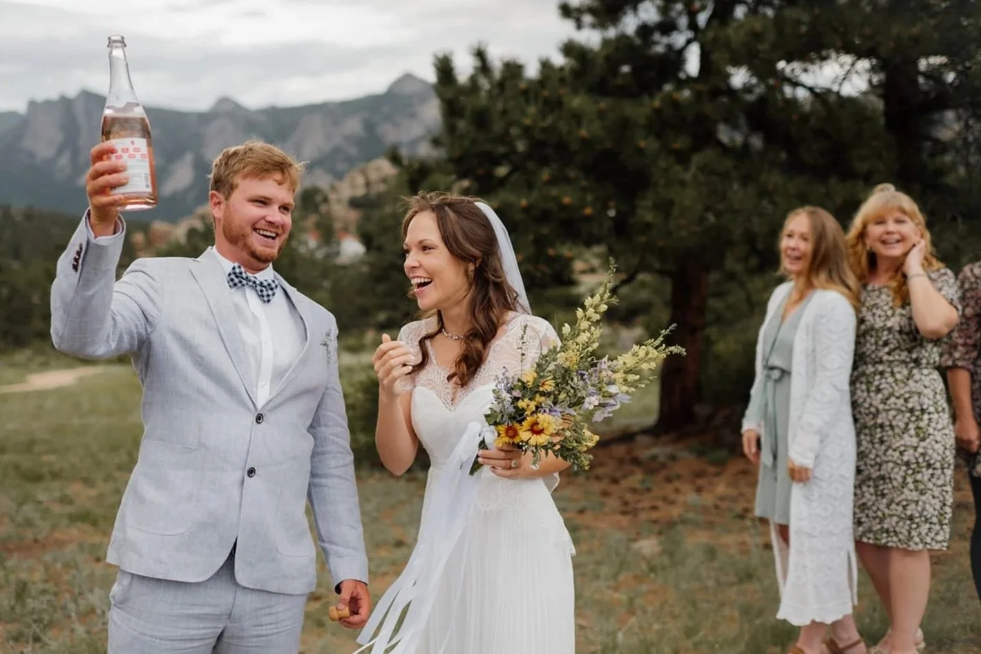 A just-married eloping couple laughs in a forest with mountain backdrop as the groom holds up a bottle of champagne while guests laugh too.