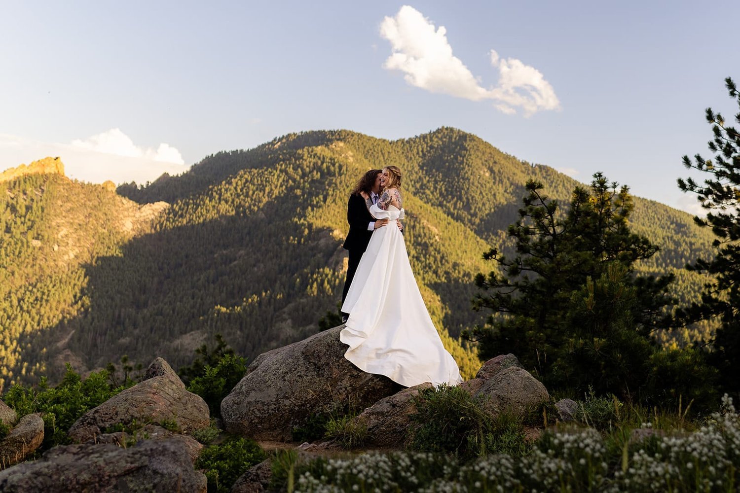 A groom kisses his bride while standing on a rock at the base of a huge green mountain behind them.